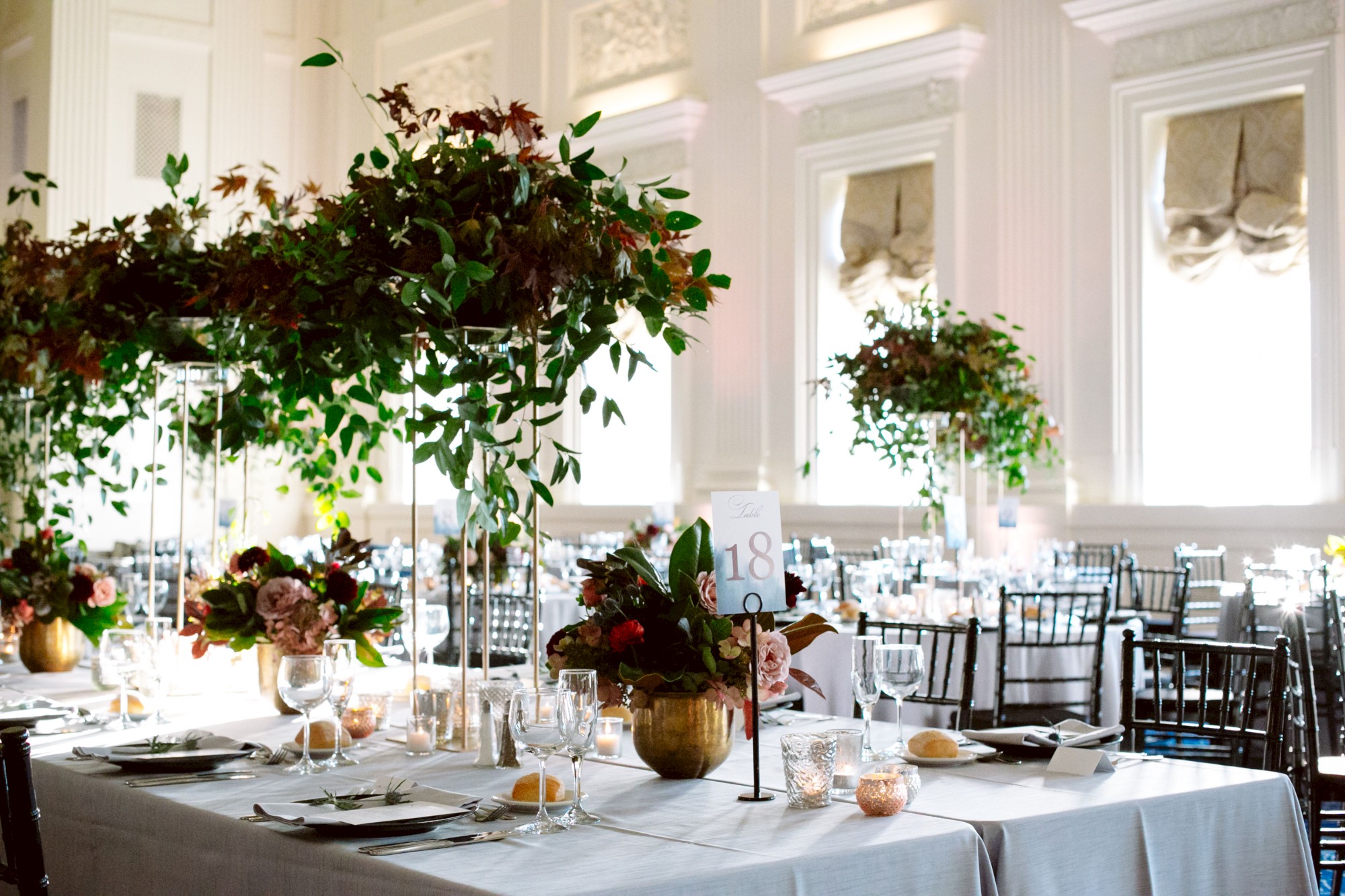A beautifully decorated banquet hall with elegantly set tables, lush floral centerpieces, and tall windows allowing natural light to pour in.