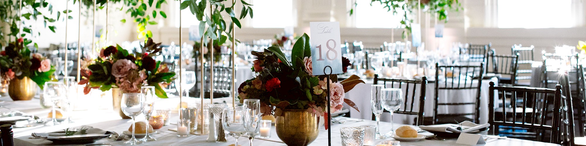 A beautifully decorated banquet hall with elegantly set tables, lush floral centerpieces, and tall windows allowing natural light to pour in.