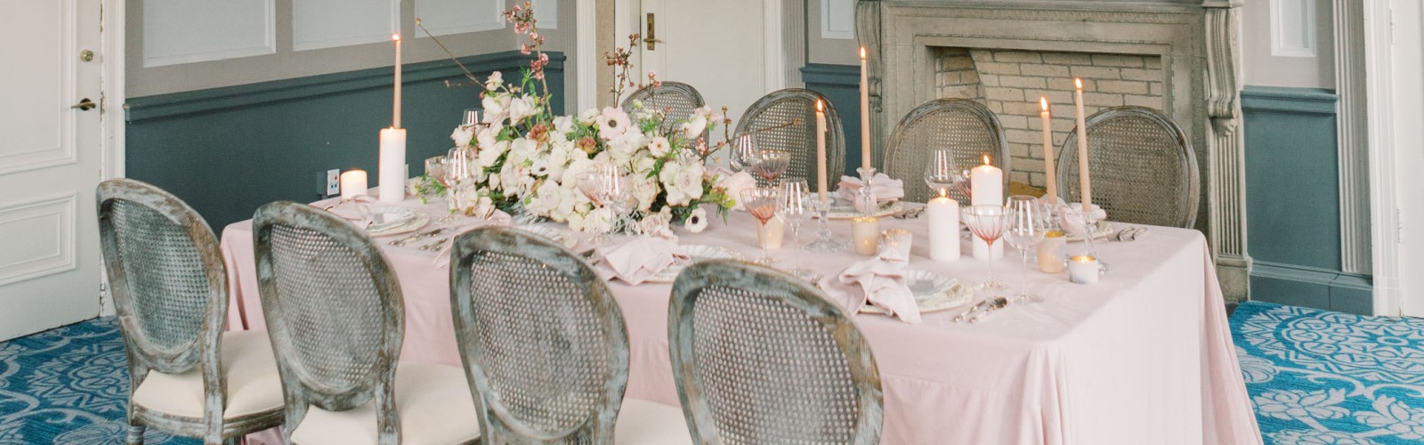 An elegant dining room with a pink tablecloth, floral centerpiece, ornate fireplace, large mirror, and chandelier, with blue patterned carpet.