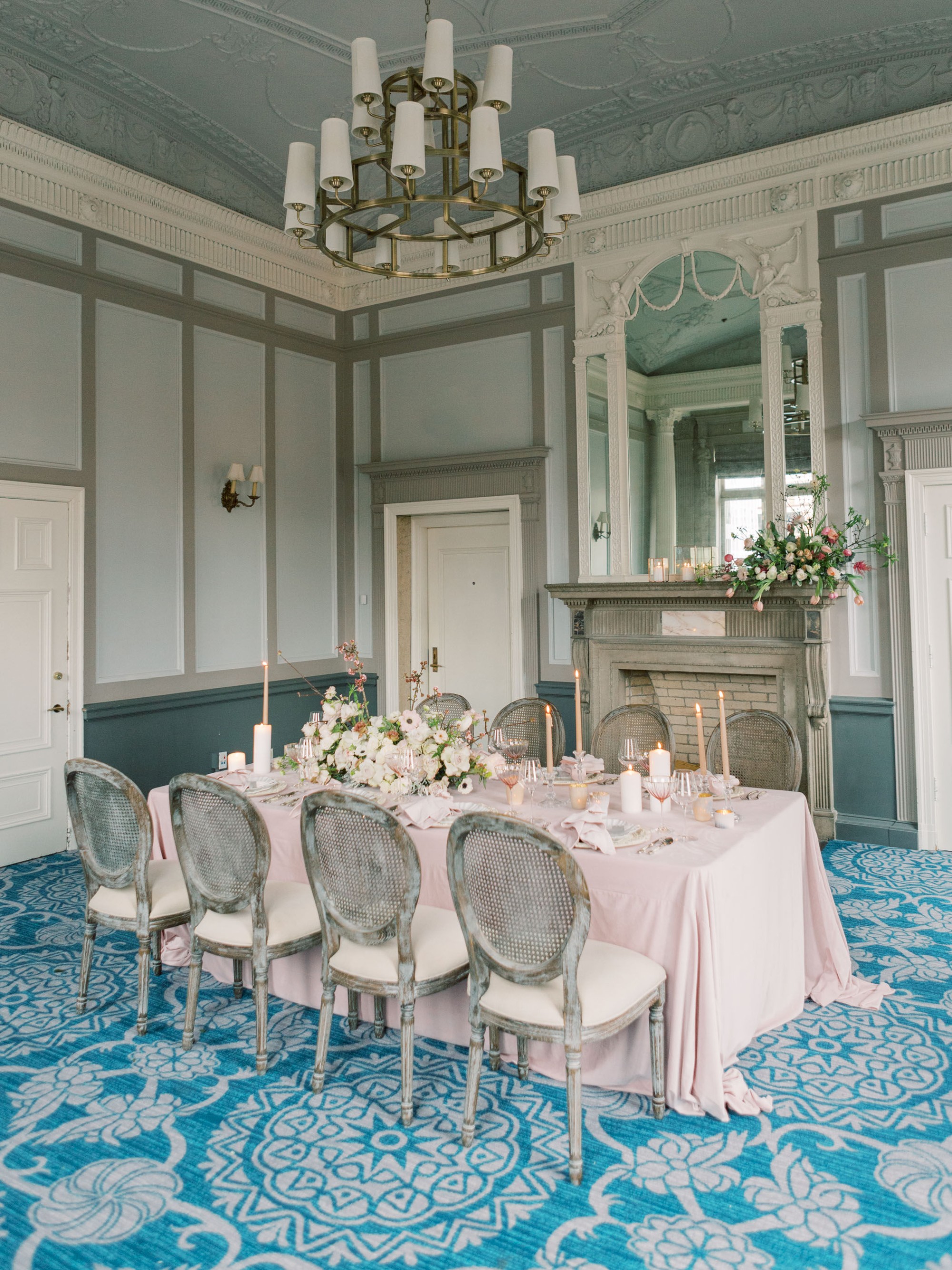 An elegant dining room with a pink tablecloth, floral centerpiece, ornate fireplace, large mirror, and chandelier, with blue patterned carpet.