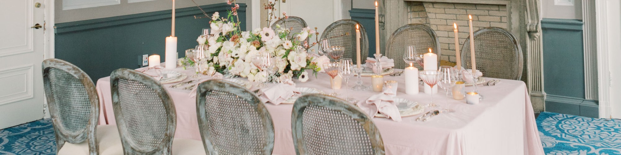 An elegant dining room with a pink tablecloth, floral centerpiece, ornate fireplace, large mirror, and chandelier, with blue patterned carpet.