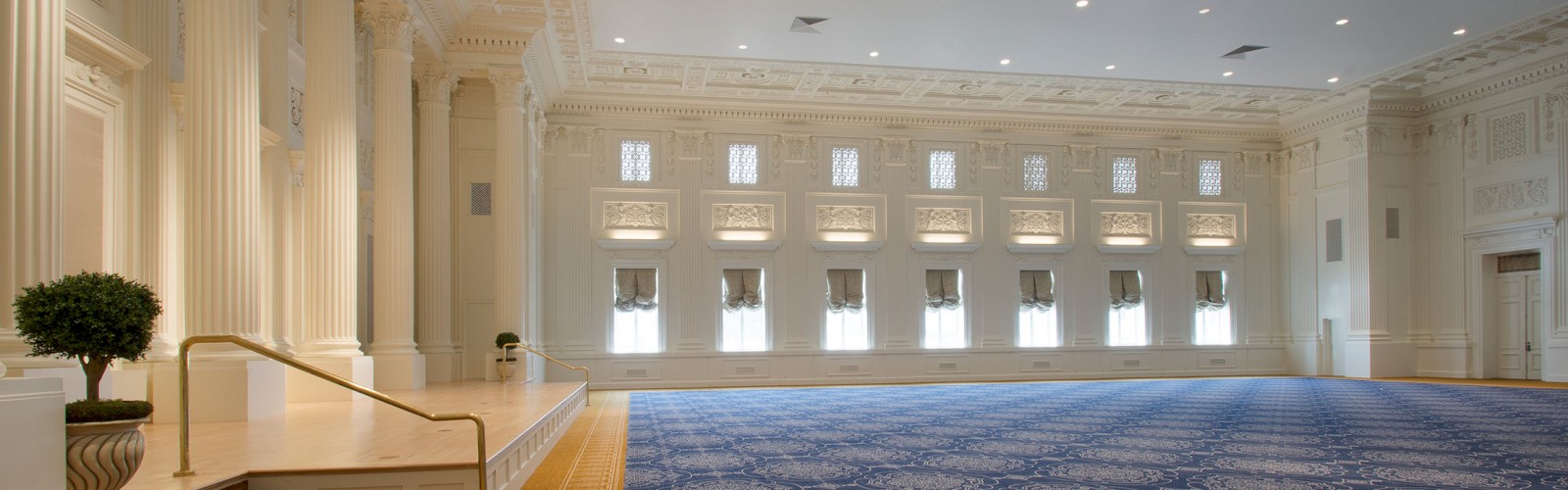 A large, elegant hall with ornate columns, decorative ceiling, and patterned carpet, features a small stage with potted plants beside it.