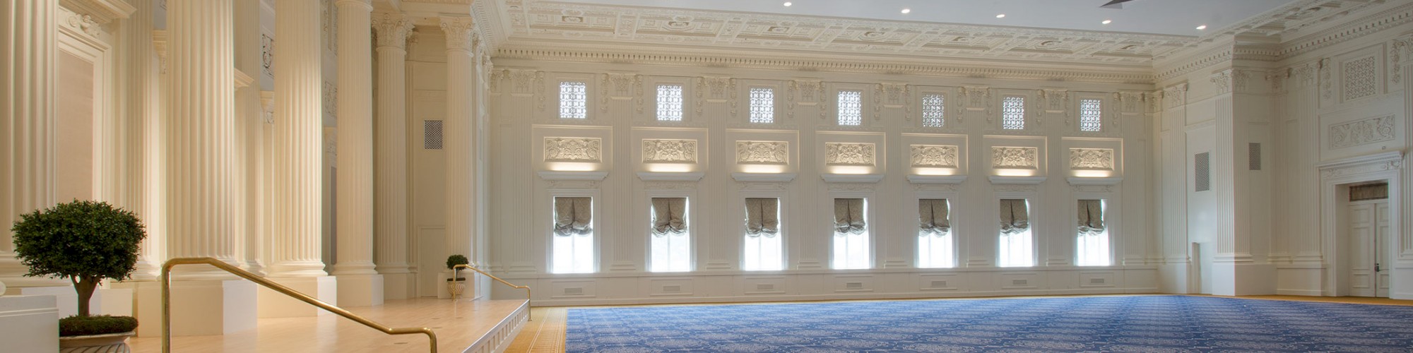 A large, elegant hall with ornate columns, decorative ceiling, and patterned carpet, features a small stage with potted plants beside it.