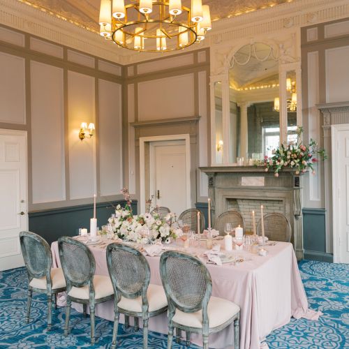 A beautifully decorated dining room with a chandelier, large mirror, flower arrangements, and a table set for a formal meal, on a blue patterned carpet.