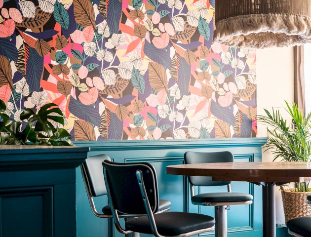 A vibrant café interior with a floral-patterned wall, teal paneling, modern chairs, wooden tables, and a fringed ceiling light fixture ending the sentence.