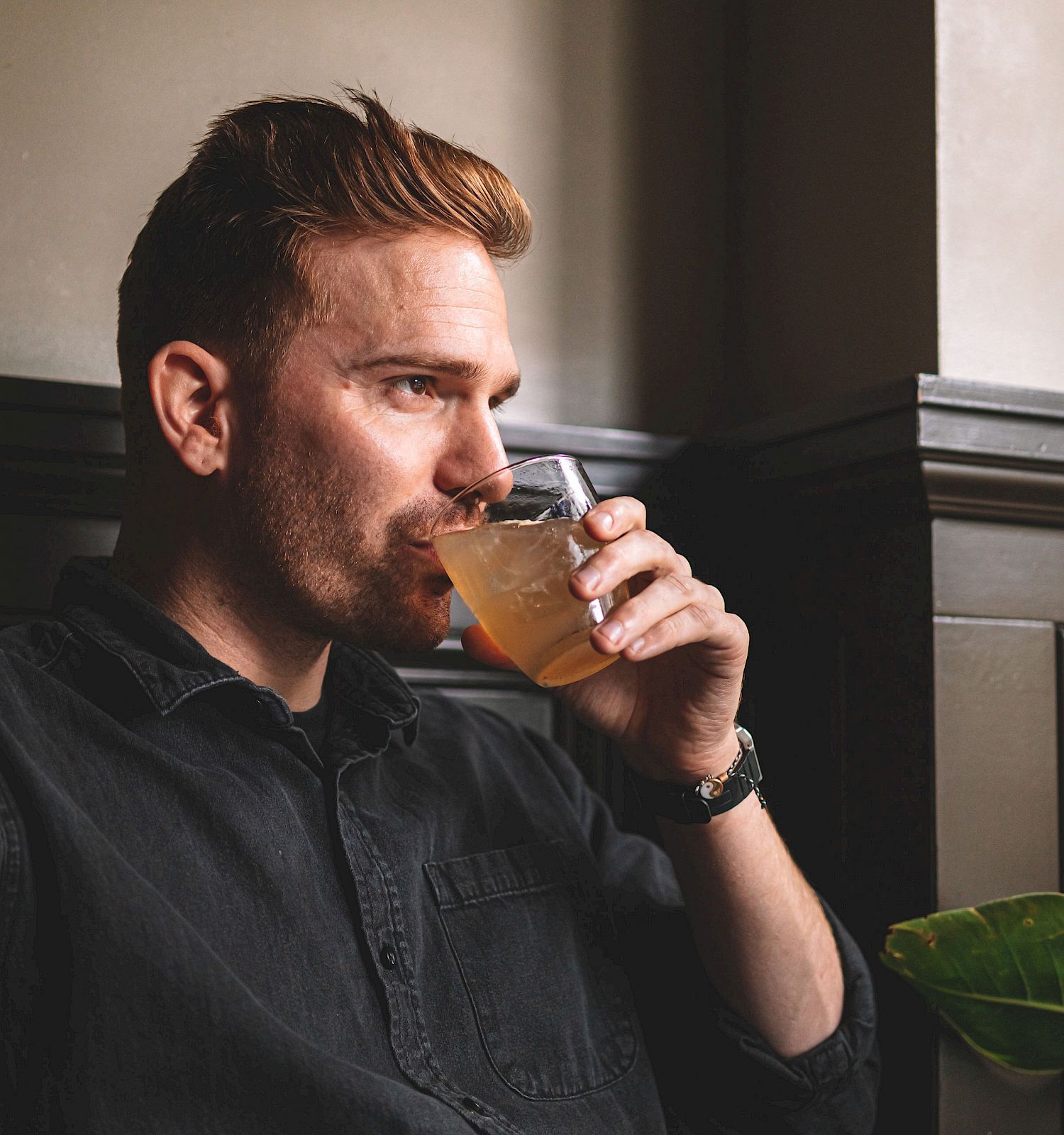 A person with short hair and a beard is seated by a window, drinking from a glass. They are wearing a dark shirt and have a calm expression.
