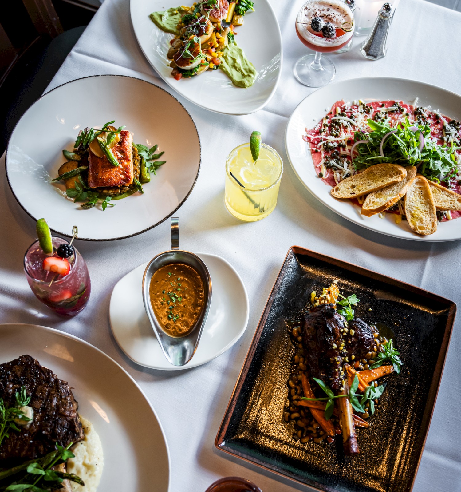 A table with a variety of gourmet dishes, including steak, pasta, and drinks, set in a fine dining atmosphere, viewed from above.