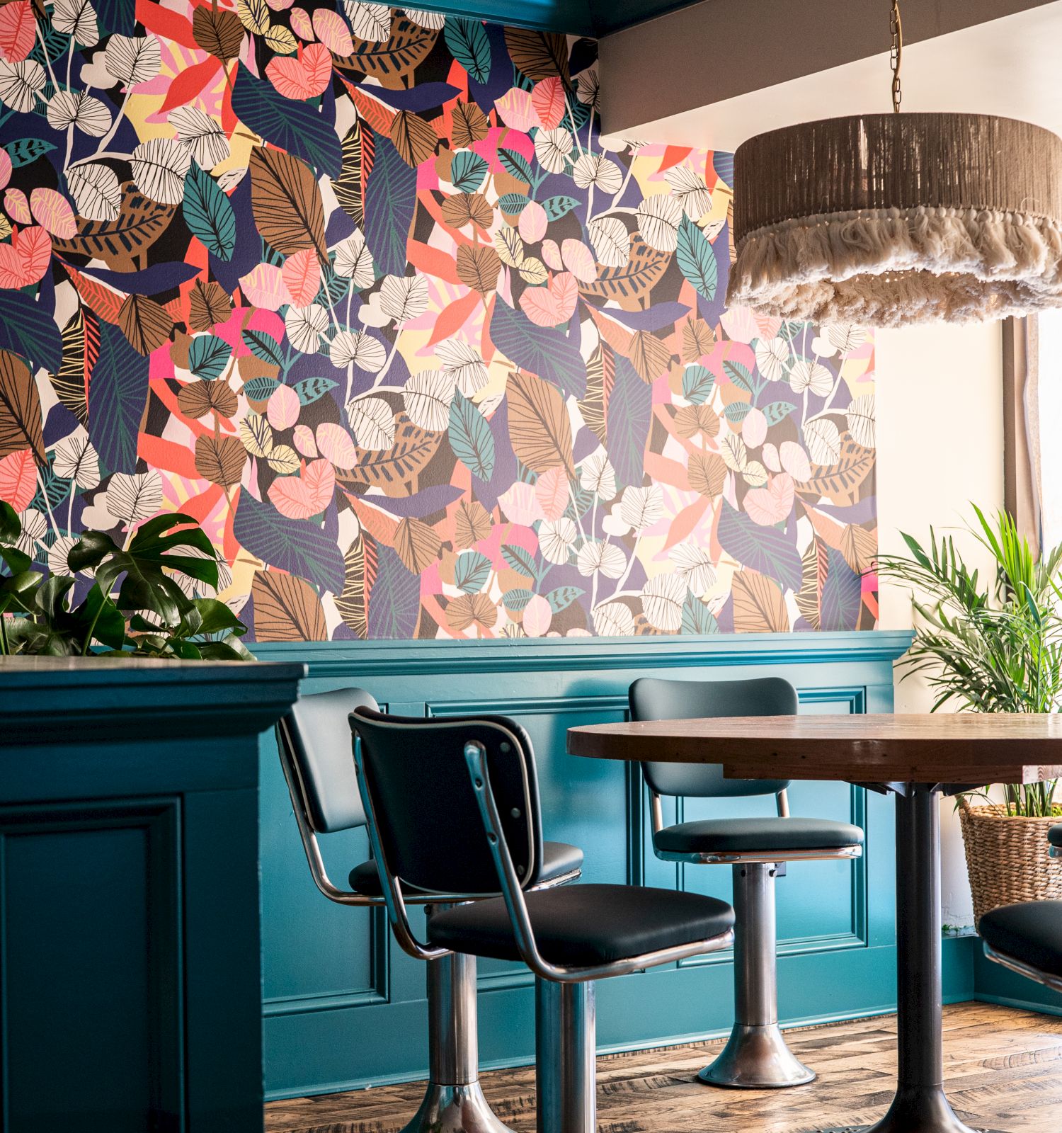 A cozy cafe with colorful floral wallpaper, teal paneling, wooden flooring, modern chairs around tables, and a funky fringe lampshade.