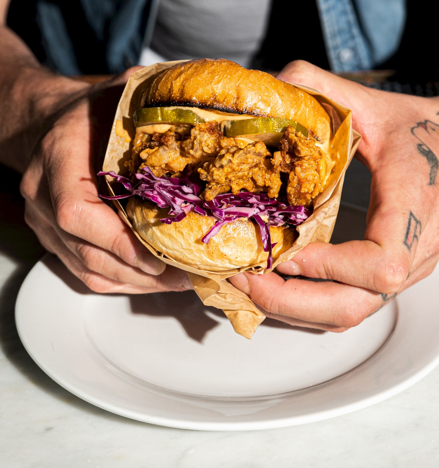 A person holds a fried chicken sandwich with pickles and purple slaw, wrapped in paper, above a white plate on a marble surface.