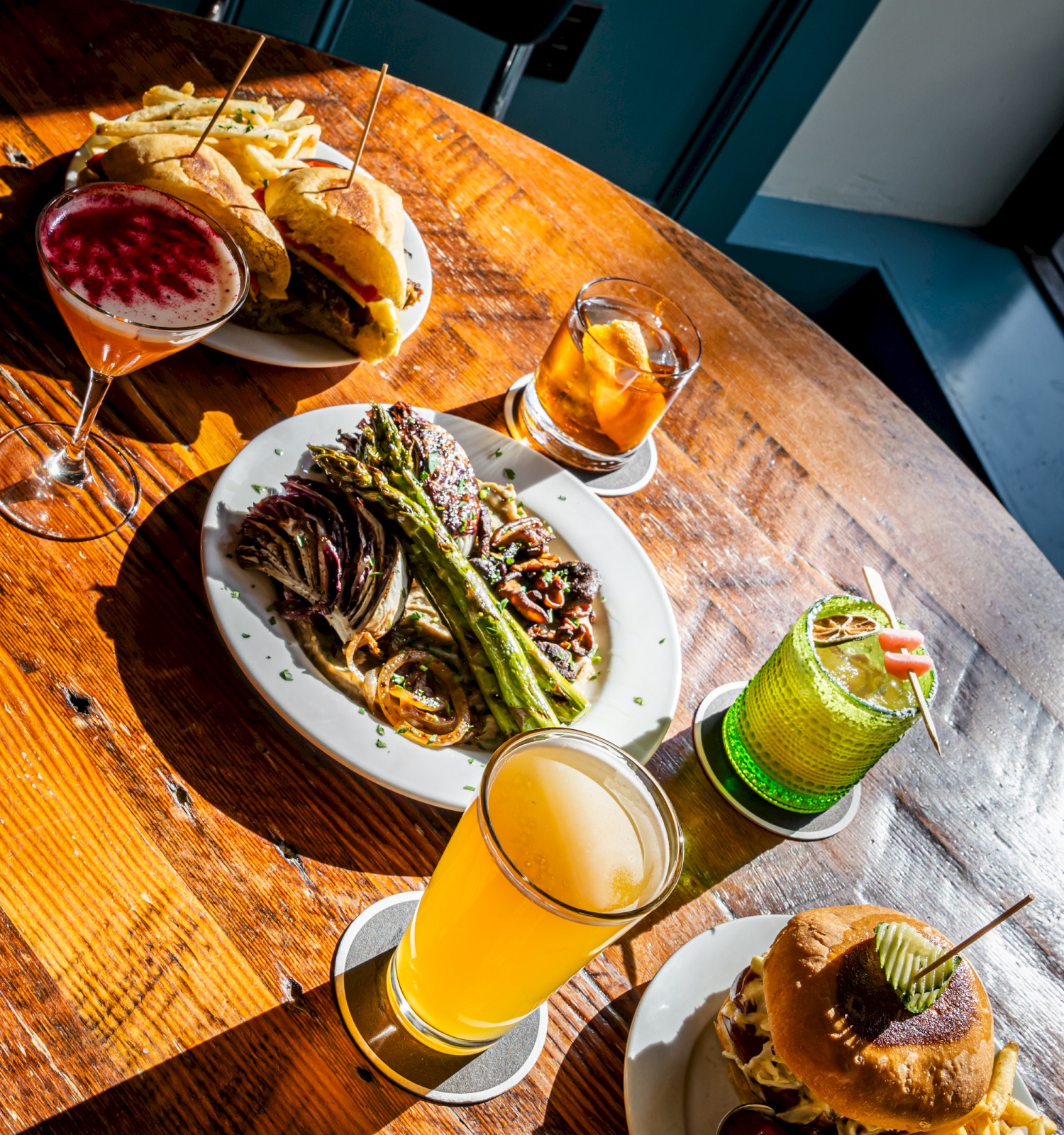 The image shows a table with burgers, fries, drinks (cocktails and beer), and a dish with a green vegetable, possibly asparagus.