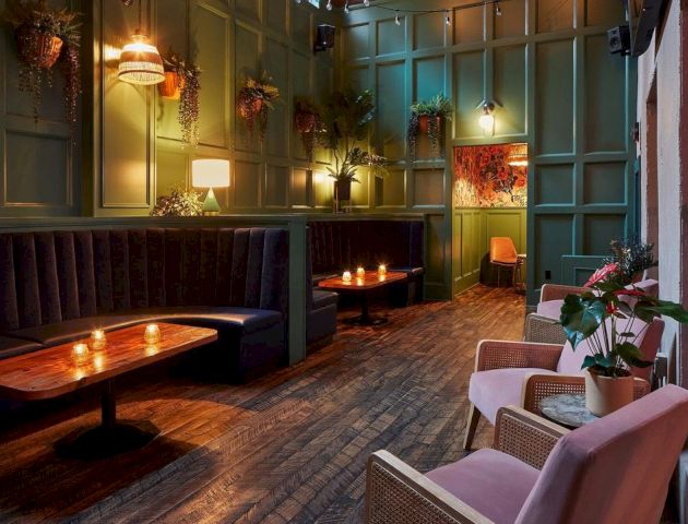 A cozy, dimly lit lounge with green paneled walls, wooden tables, cushioned seating, candles, potted plants, and decorative wall art.