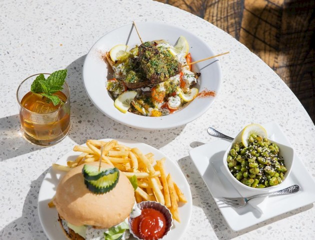 A table with a burger and fries, a bowl of edamame, a salad-like dish with skewers, an iced drink with mint, and a lemon garnish.