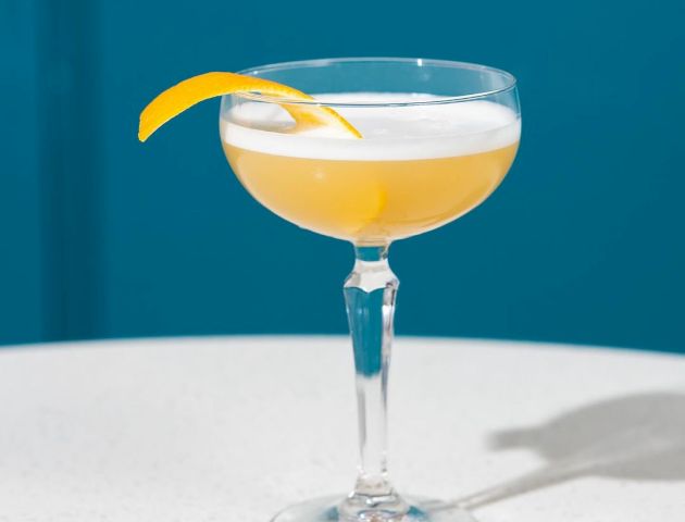 The image shows a cocktail in a coupe glass, garnished with a twist of lemon peel, placed on a white surface against a blue background.