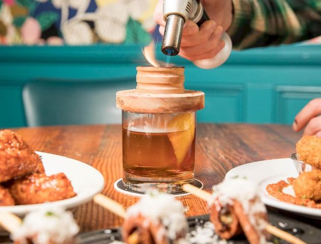 A hand is using a torch on a cocktail jar with dishes of appetizers around it on a wooden table, in a vibrant, colorful restaurant setting.