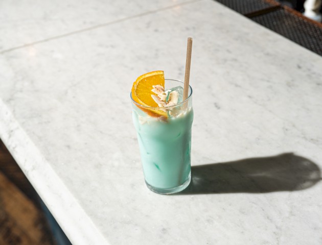 A glass filled with a blue drink, garnished with an orange slice and straw, placed on a marble surface.