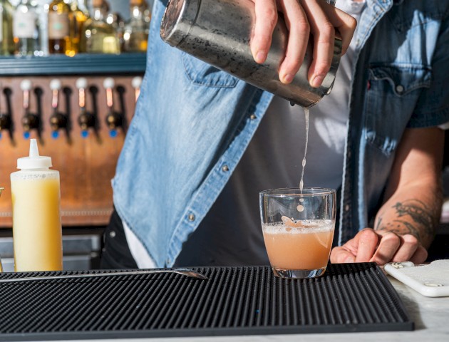 A person is pouring a drink from a shaker into a glass at a bar, with bottles in the background and a squeeze bottle nearby.