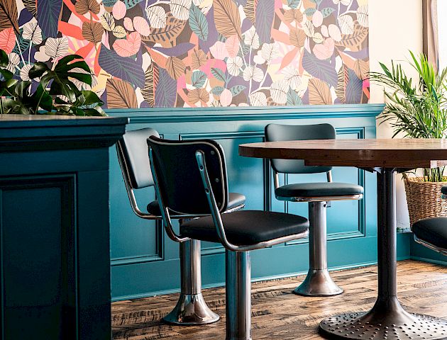 A stylish room featuring a vibrant floral wall, teal paneling, modern chairs with a round table, and a fringed pendant lamp.