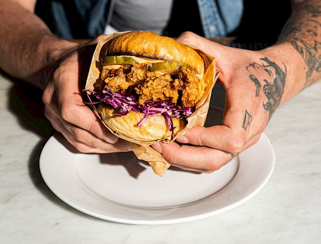 A person holding a fried chicken sandwich with pickles and coleslaw on a plate, showing their tattoos.
