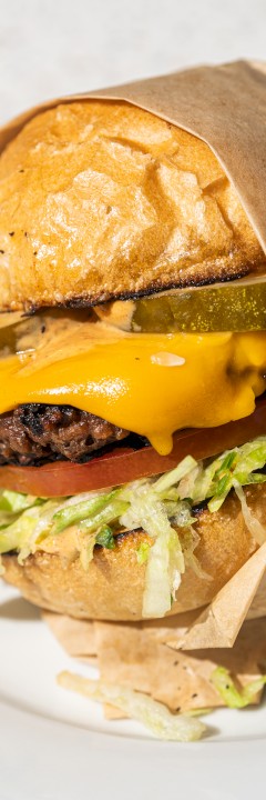 A cheeseburger with lettuce, tomato, pickles, and cheese in a bun, partially wrapped in paper, served on a white plate.