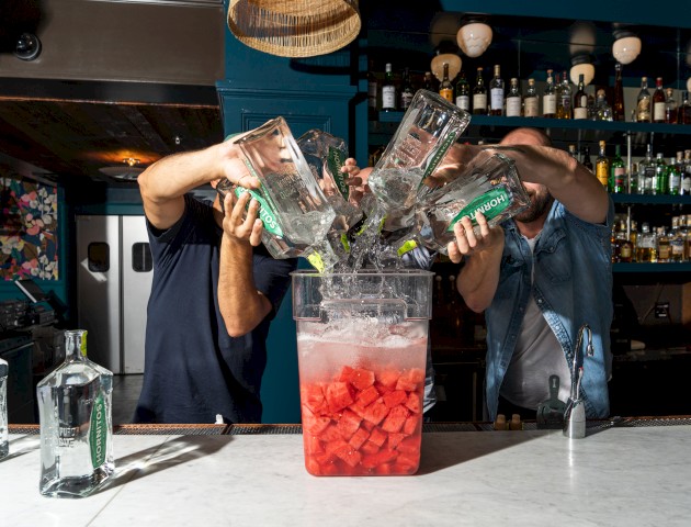 Three people pouring bottles of alcohol into a large container filled with ice and chopped watermelon at a bar counter.