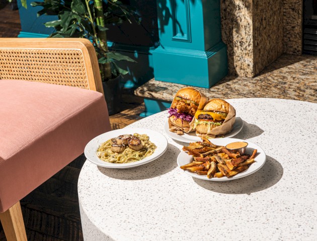 A round white table with various plates of food, including a burger, chips, and a green dish, next to a pink chair and a plant.