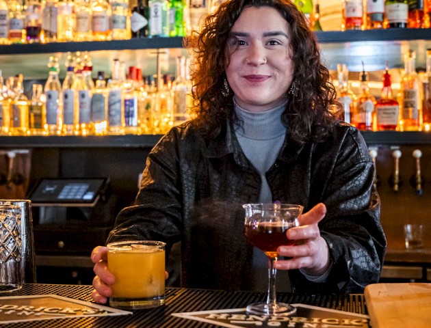 A bartender standing behind a counter holding two drinks, with shelves of liquor bottles in the background.