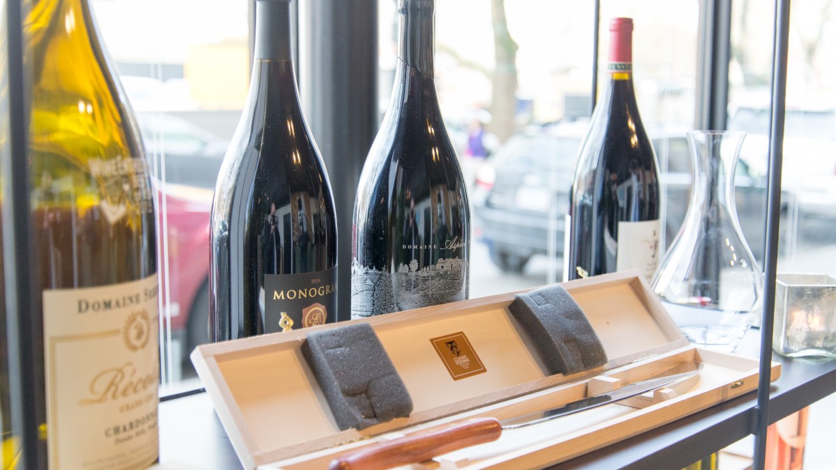 The image shows a display of wine bottles and a wine opener set inside a store, with large windows revealing a parking lot outside.