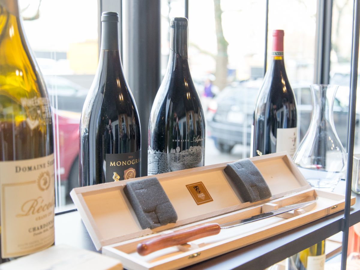 The image shows a display of wine bottles and a wine opener set inside a store, with large windows revealing a parking lot outside.