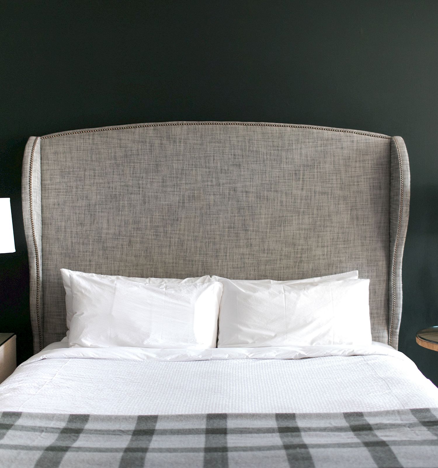 A neatly made bed with a large padded headboard, flanked by two bedside tables each with a lamp, a small clock, and a green decorative item.