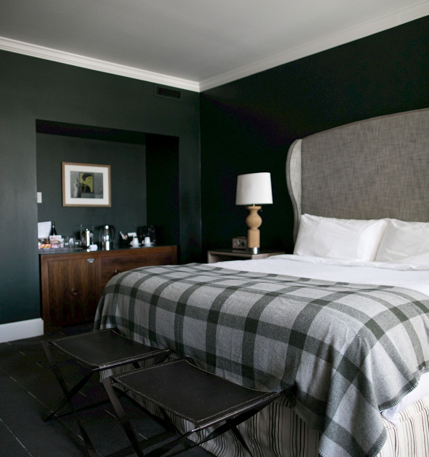 A stylish bedroom features a large bed with a plaid blanket, two stools at the foot, a nightstand with a lamp, and a counter with a framed picture.