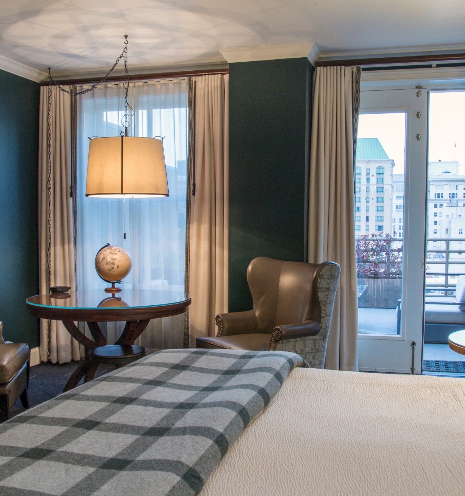 A cozy hotel room with a plaid blanket on the bed, two armchairs, a table with a globe, large windows, and a balcony view of buildings outside.