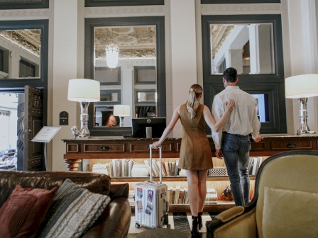 A couple stands in a stylishly decorated room with mirrors, lamps, and a cozy sofa, looking at something off-camera while holding a suitcase.