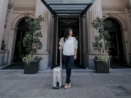 A person stands with luggage in front of an ornate building labeled 