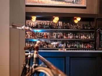 A bar with a fully stocked shelf, topped with a painting of an old car scene. A bicycle is visible in the foreground.