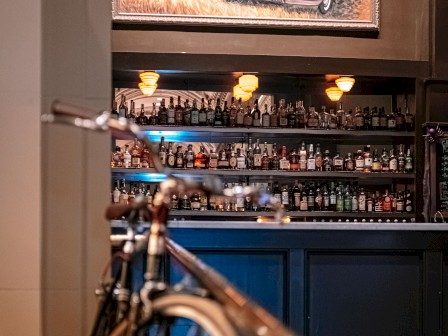 A bar with a fully stocked shelf, topped with a painting of an old car scene. A bicycle is visible in the foreground.