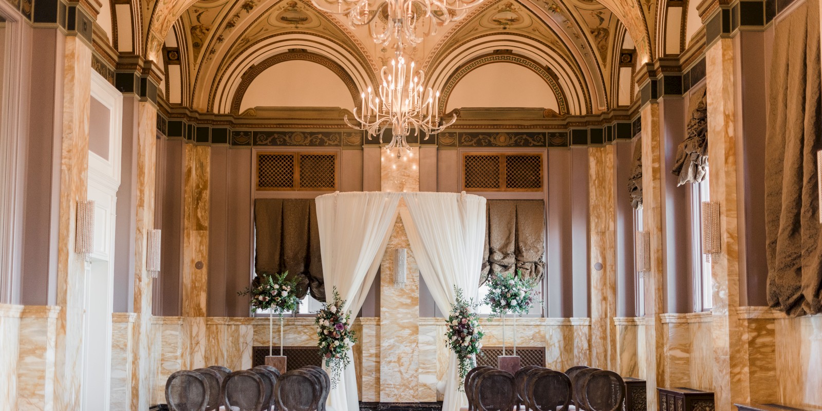 An elegant room set for a wedding ceremony with a decorative arch, floral arrangements, chairs, and ornate chandeliers under a vaulted ceiling.