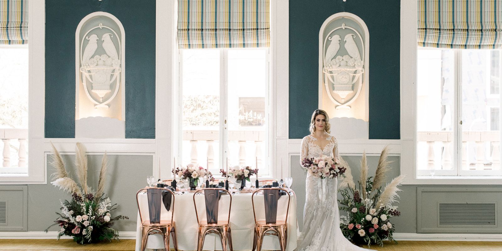 A bride in a wedding dress stands beside an elegantly set table with floral arrangements, in a room with ornate decor and large windows.