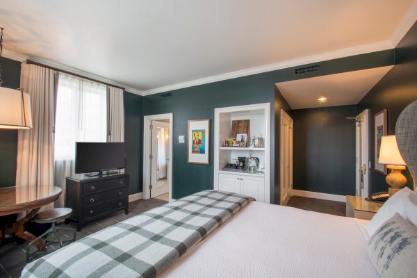 A cozy bedroom with a plaid blanket, dark green walls, TV on a dresser, a side table, and a small closet with amenities.