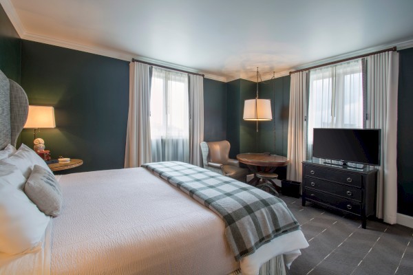 A cozy hotel room with a double bed, checkered blanket, armchair, round table, lamp, TV on dresser, and curtains over two windows.