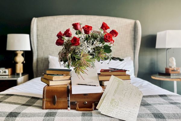 A bouquet of red roses, books, and a vintage suitcase are arranged on a bed with plaid bedding in a cozy room with lamps on side tables.