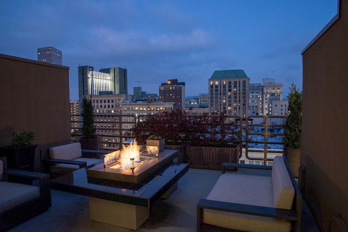 A rooftop patio with seating around a lit fire pit overlooks a city skyline at dusk, capturing the serene ambiance of an urban evening.