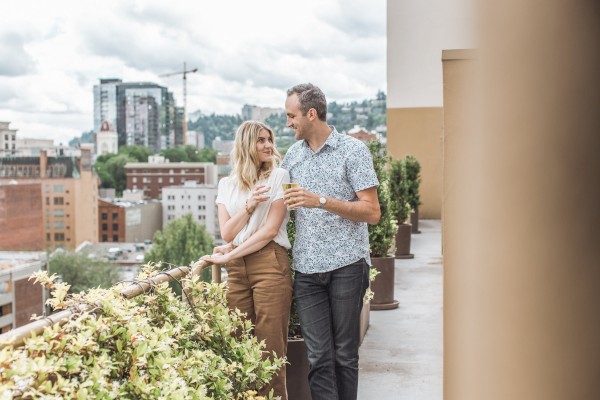 A couple stands on a balcony overlooking a city, surrounded by plants, with buildings and a cloudy sky in the background.