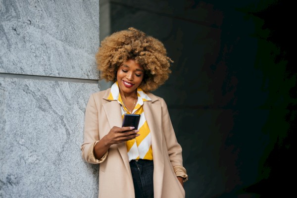 A person with curly hair is smiling and looking at their phone while leaning against a wall. They are wearing a beige coat and a yellow striped shirt.