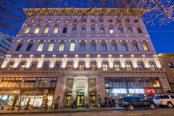 A historic multi-story building with ground-floor shops and illuminated windows at night. Cars are parked on the street in front of it.