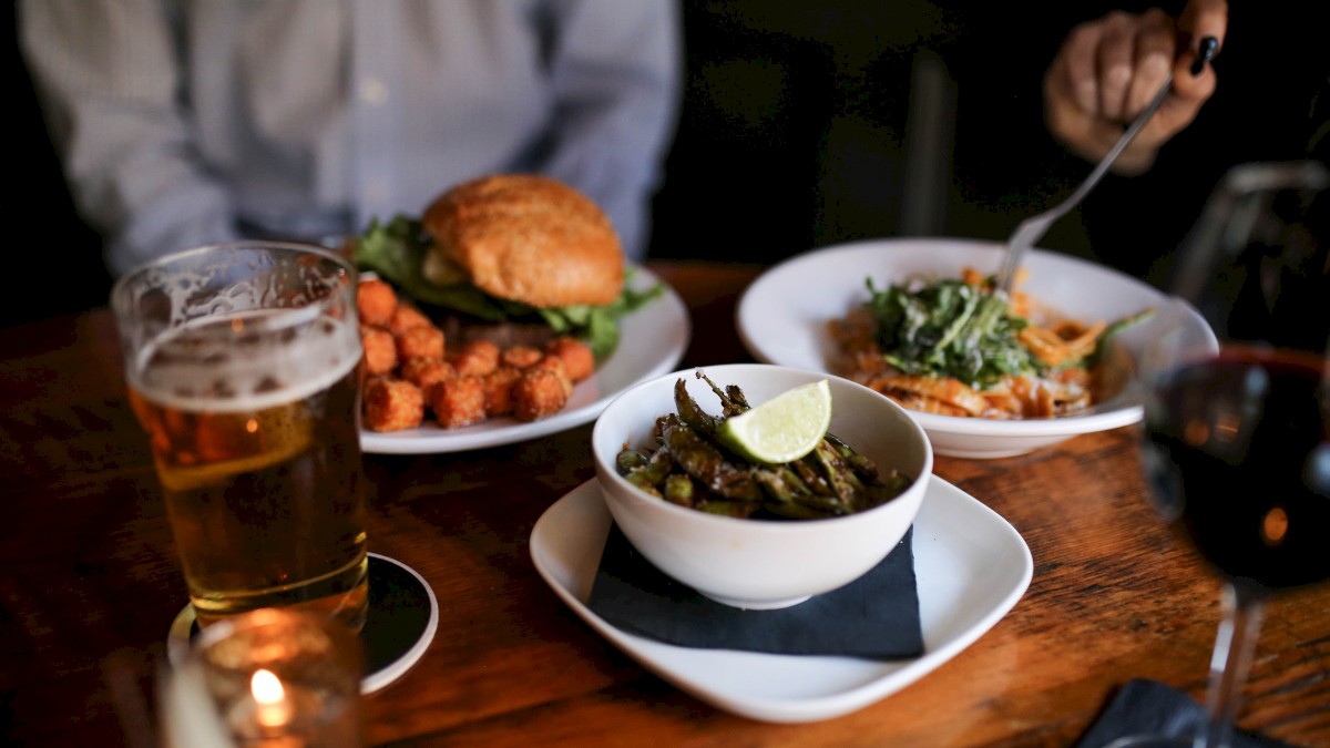 A table with two people dining, featuring a burger with tater tots, a salad, a bowl of lime-topped dish, a beer, and a glass of red wine.