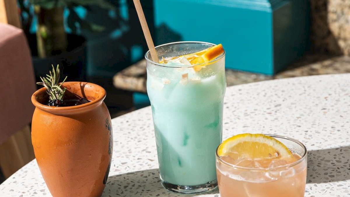 The image shows a terracotta pot with a plant, a blue drink with an orange garnish, and a clear drink with an orange slice on a white table.