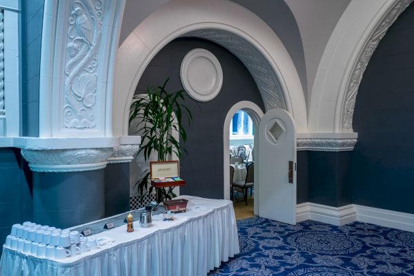 The image shows a decorative room setup for a gathering with a table of refreshments, arched doorways, ornate walls, and a blue carpet.