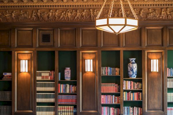 A well-lit, elegant wooden bookshelf lined with books, decorative vases, and ornate ceiling details with a chandelier hanging from above.