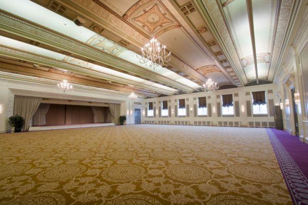The image shows an elegant, spacious ballroom with chandeliers, ornate ceiling, patterned carpet, and a stage area, designed for large events and gatherings.