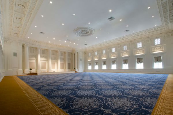 The image shows a large, empty hall with ornate ceilings and chandeliers. The hall has blue and gold carpeting with intricate patterns, and tall windows.